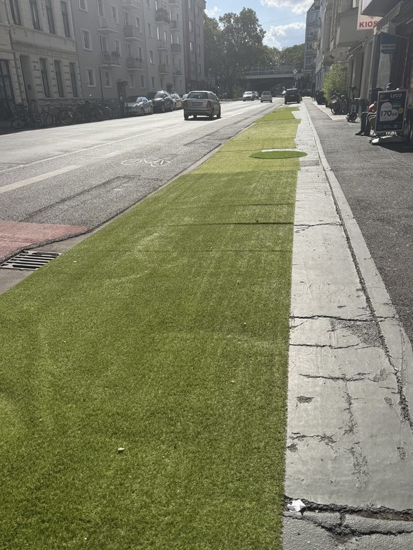 Lindenstraße stadtauswärts mit Blick zur Bahnunterführung: Neben dem Radfahrstreifen liegt ein breiter Streifen Kunstrasen auf dem Gehweg. Kein Auto steht auf dem Gehweg.